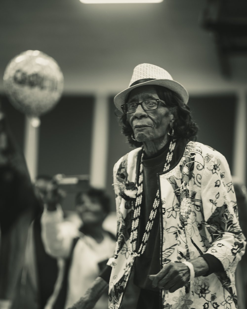 A Black Creole elder woman in a white sun hat and floral jacket standing at a community gathering