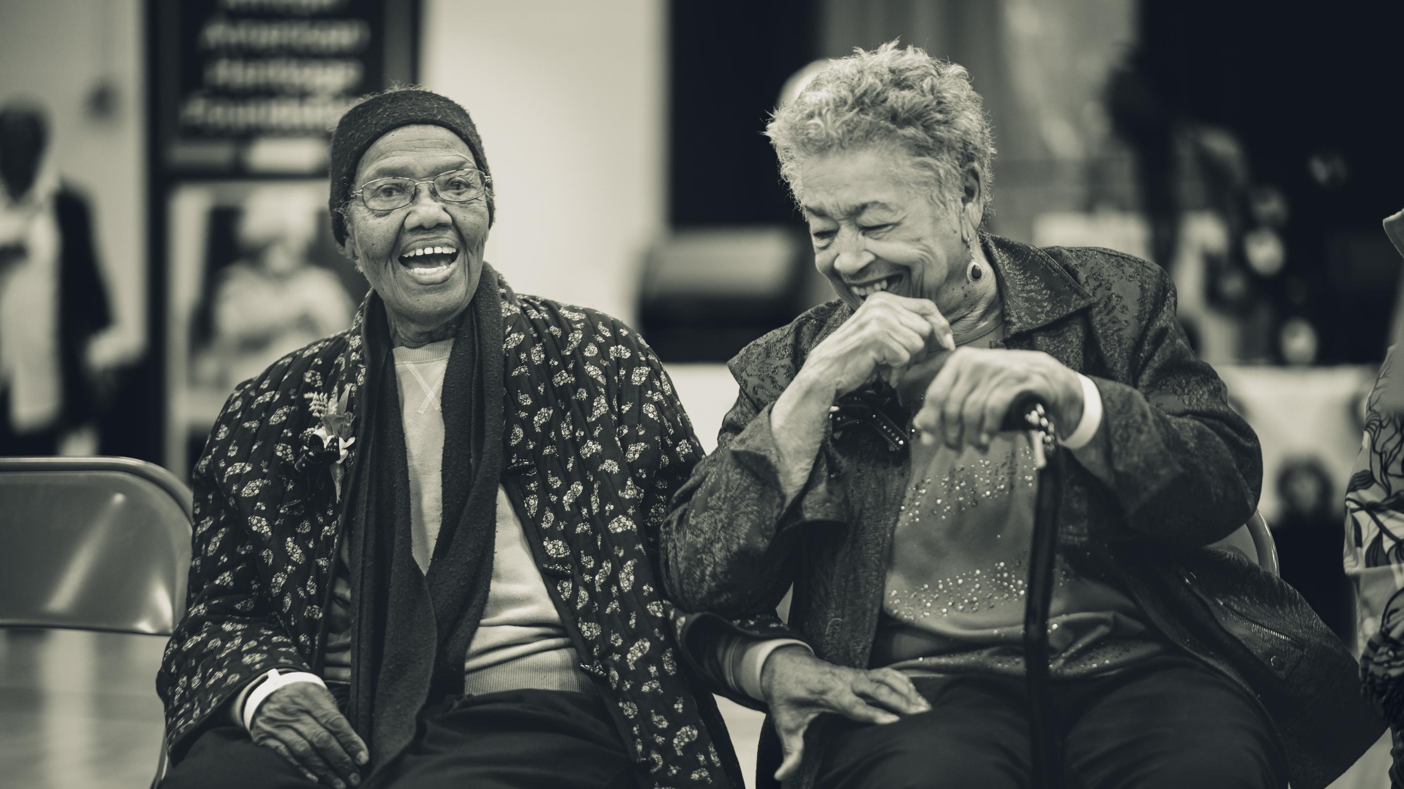 Two Black Creole elders seated side by side, laughing together at an African American Heritage Foundation gathering in Louisiana